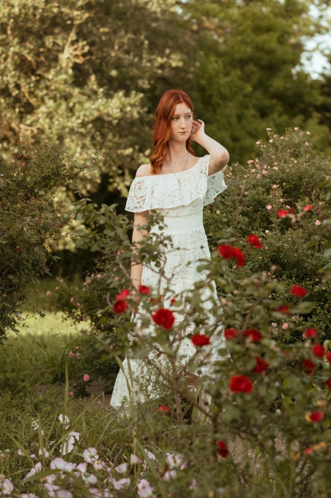 Redhead woman in white off-shoulder lace dress standing among blooming red roses in a lush garden.