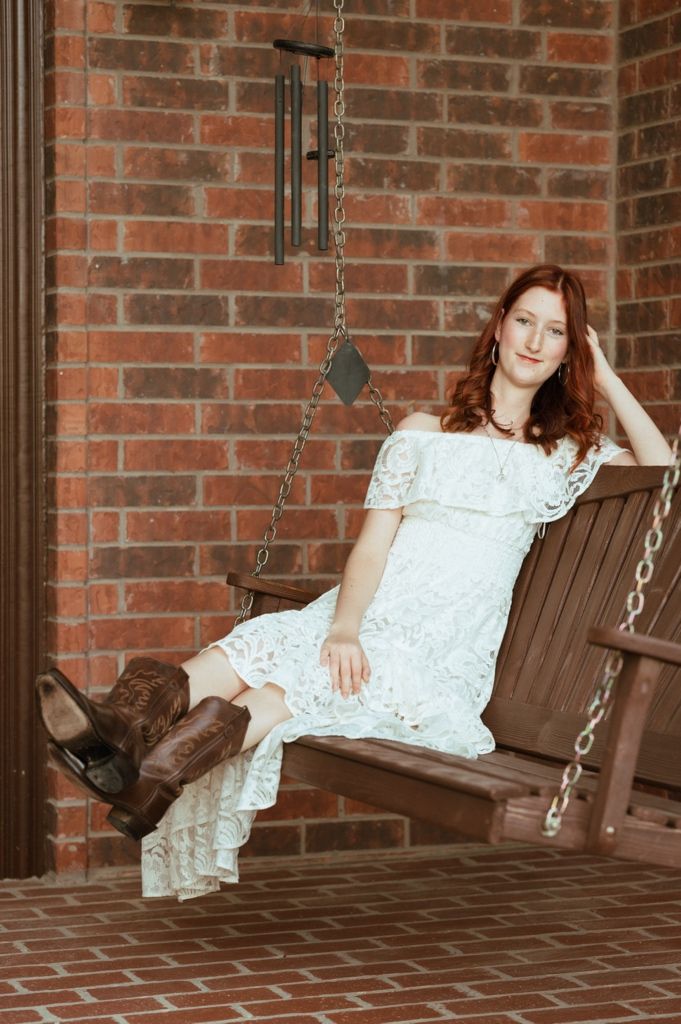 Young woman with red hair in white lace dress and cowboy boots sitting on a wooden porch swing.