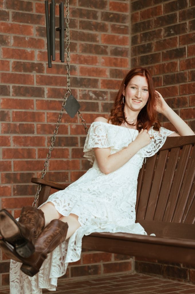 Young redhead woman in white lace dress and cowboy boots sitting on a wooden porch swing against brick wall.