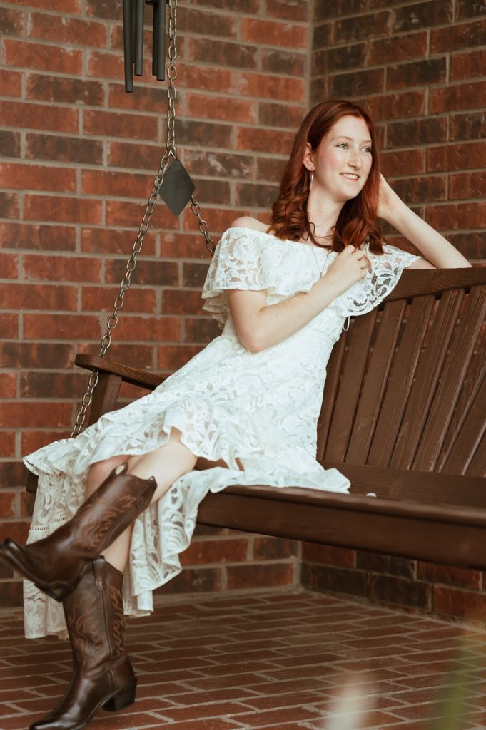 Young redhead woman in white lace dress and cowboy boots sitting on a wooden porch swing against brick wall.