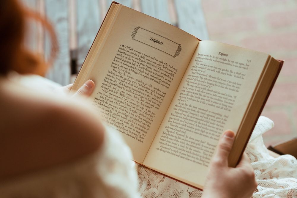 Woman with red hair reading an old vintage book, viewed from behind, holding open pages with worn edges.