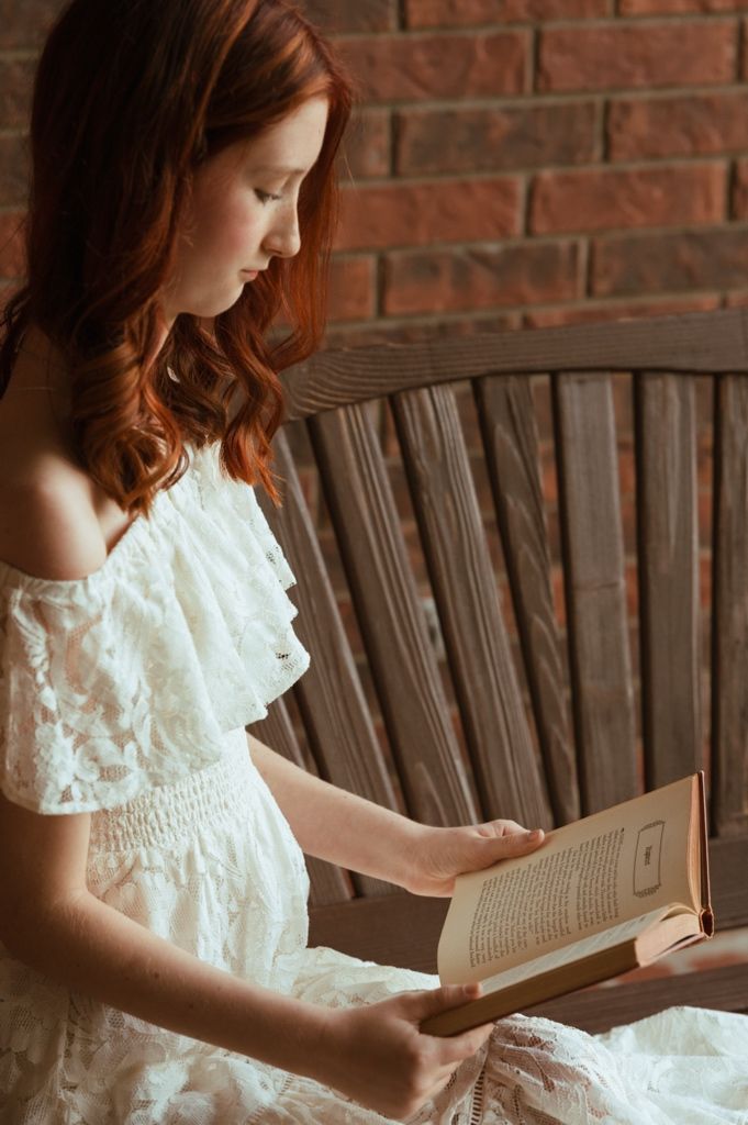 A young redhead woman in a white off-shoulder dress reads a book on a wooden bench by a brick wall.