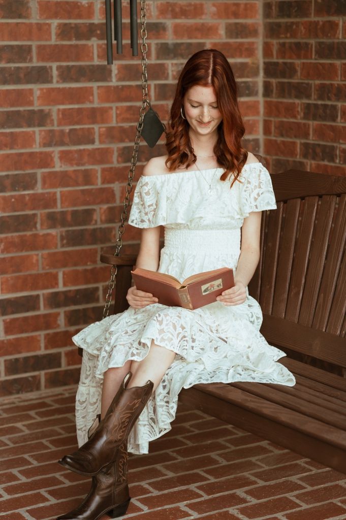 Young redhead woman in white lace dress and cowboy boots reading a book on a wooden porch swing.