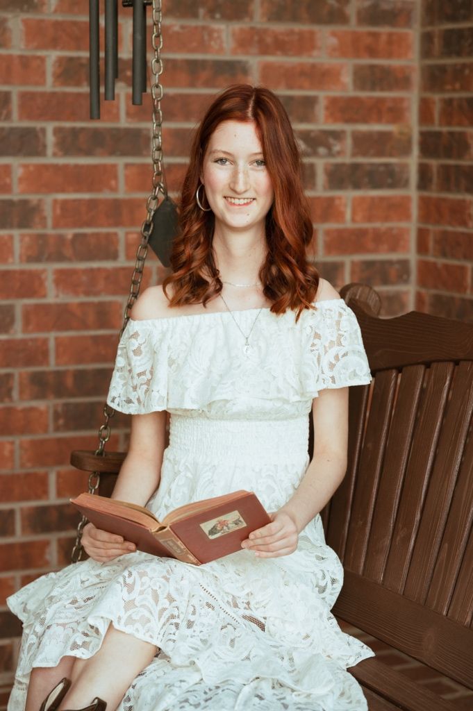 Young redhead woman in white lace off-shoulder dress sitting on bench swing holding book against brick wall.