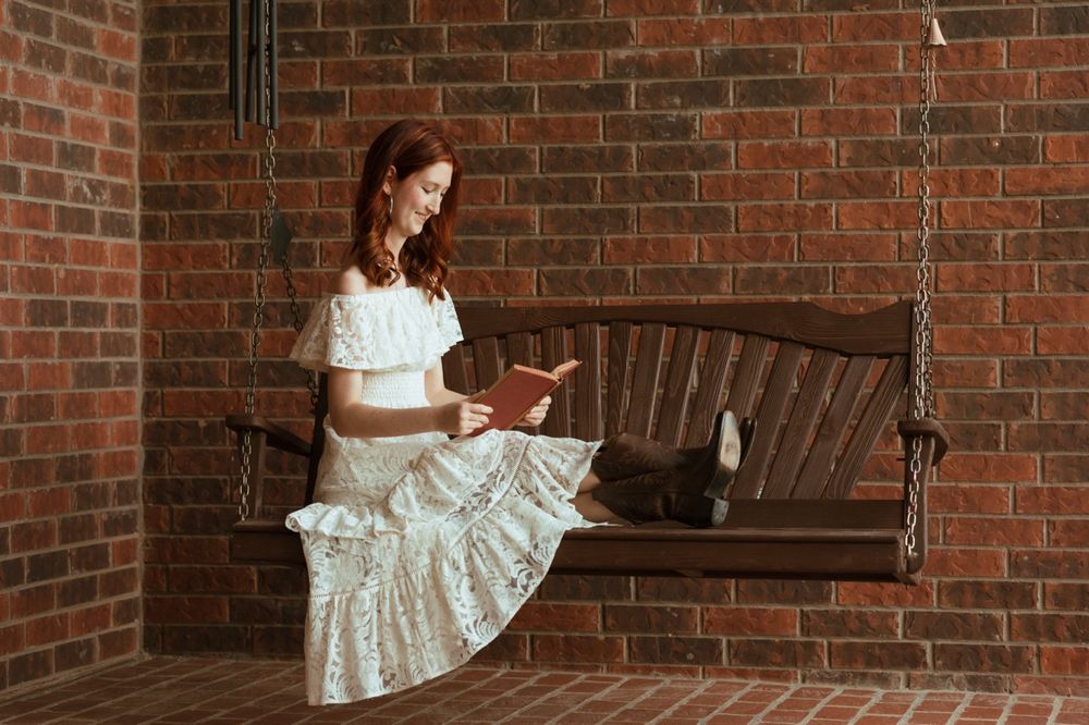 Young woman in white dress reading a book on a wooden swing bench against a brick wall.