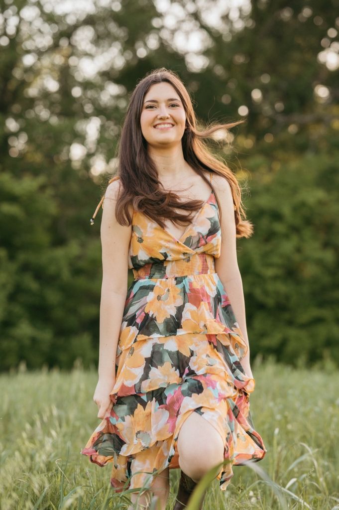 Smiling young woman in yellow floral sundress standing in green meadow with trees in background.