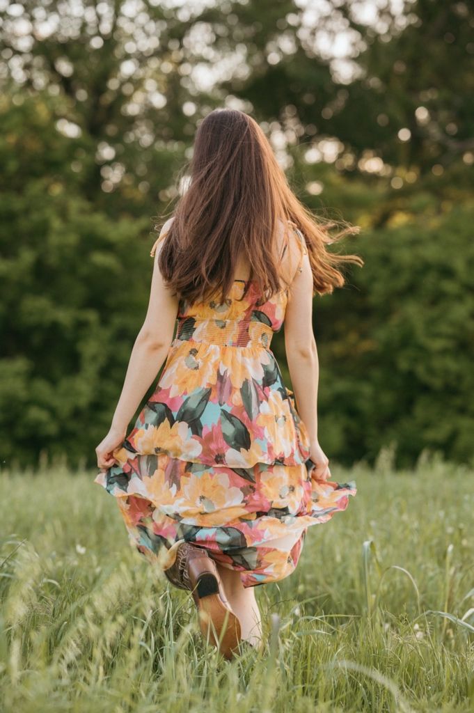 Woman in floral dress walking through green meadow, viewed from behind, long brown hair flowing in breeze.