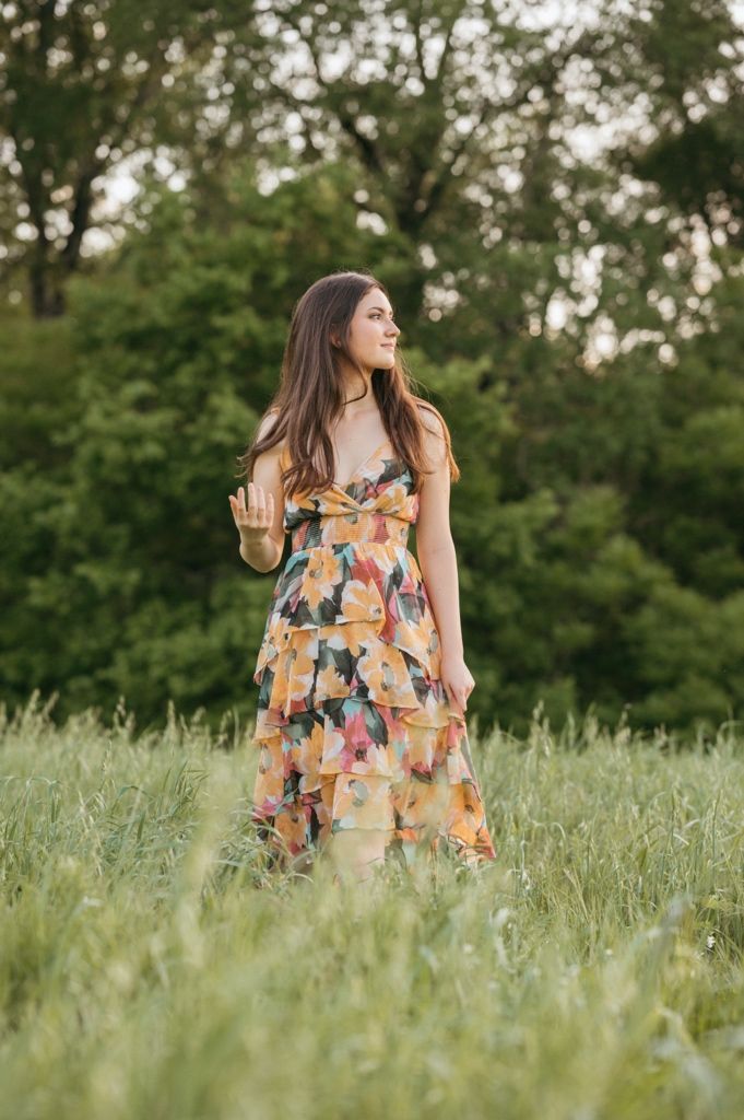 Young woman in floral tiered dress standing in lush green field surrounded by trees at golden hour.