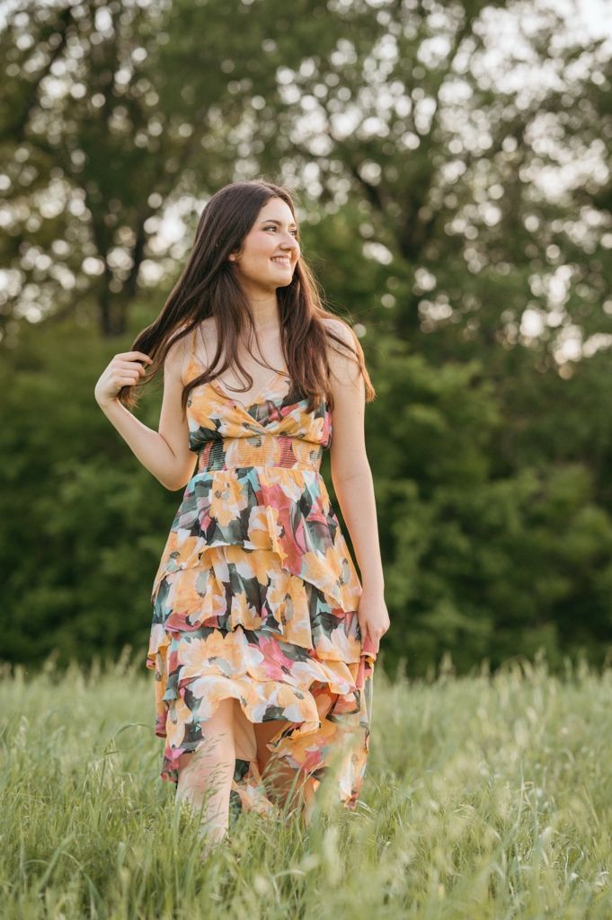 Young woman in floral ruffle dress standing in green meadow, smiling and looking away, with trees in background.