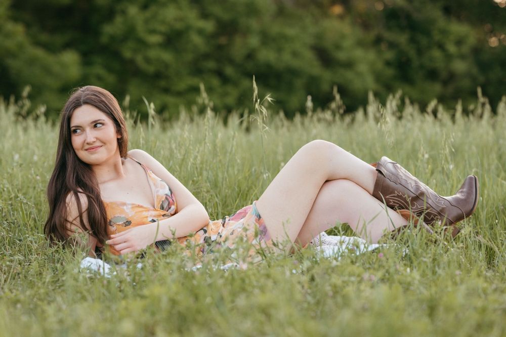 Young woman in floral dress and cowboy boots relaxing in lush green meadow grass on sunny day.