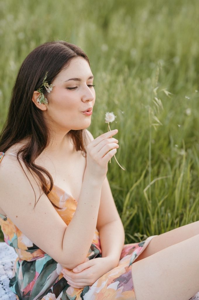 Young woman in floral dress sitting in green field, blowing dandelion seeds with eyes closed.