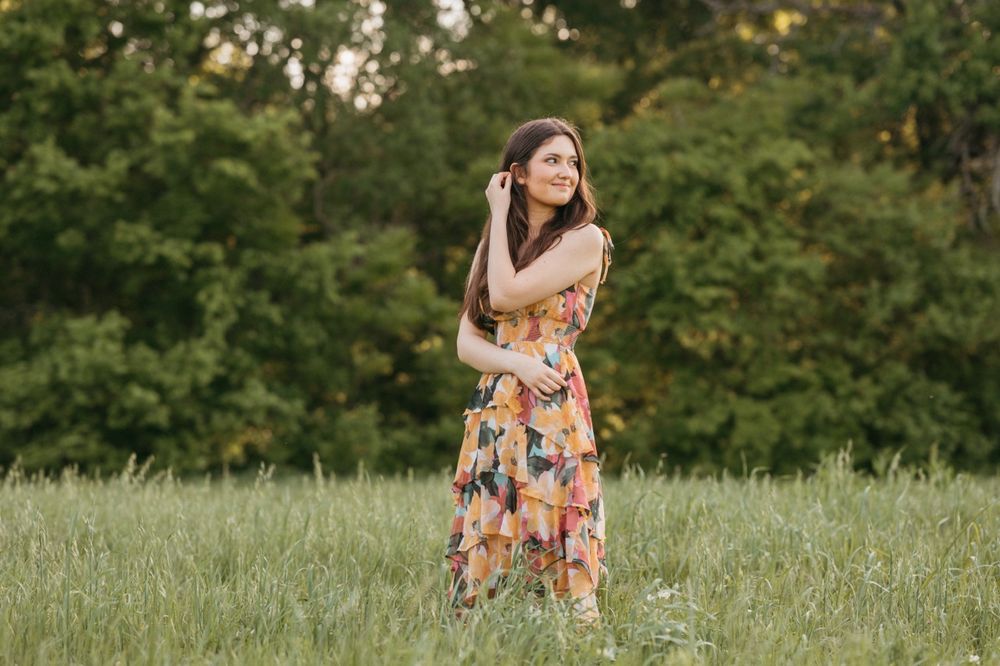 Young woman in floral maxi dress standing in green meadow with trees, posing outdoors during golden hour.