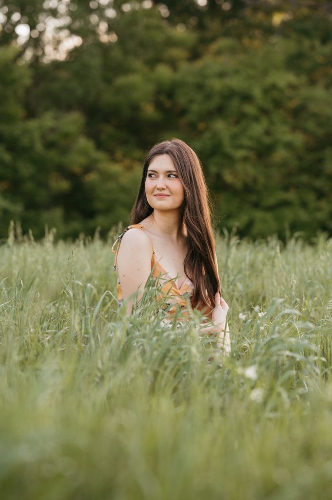 Young woman with long brown hair sitting in tall green grass field wearing floral dress, trees in background.