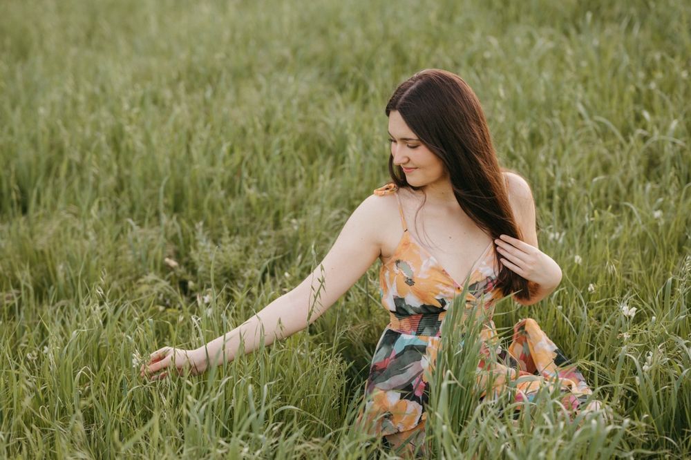Young woman in floral dress sitting in tall green grass field, smiling softly while touching plants on a serene day.