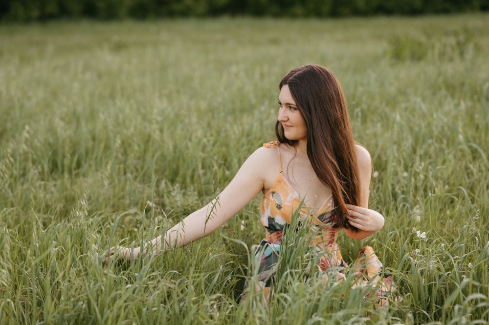 Young woman in floral dress sitting in tall green grass field, looking sideways with long brown hair.