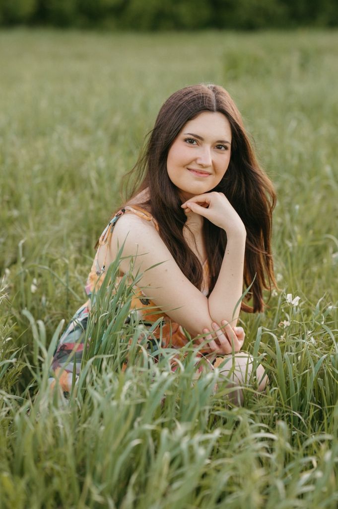 Young woman with long brown hair sitting in a lush green field, smiling warmly at the camera.