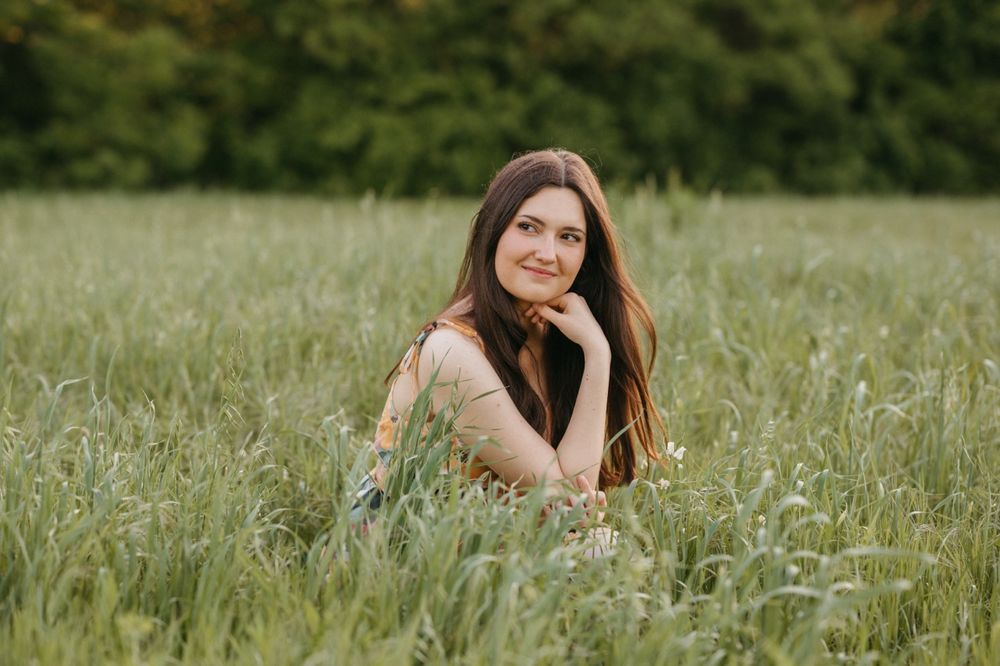 Smiling brunette woman sitting in lush green grass field, resting chin on hand in natural setting.