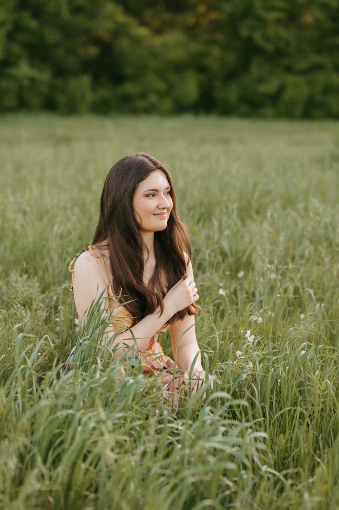 Young woman with long brown hair sitting in tall green grass field, smiling softly in warm natural light.