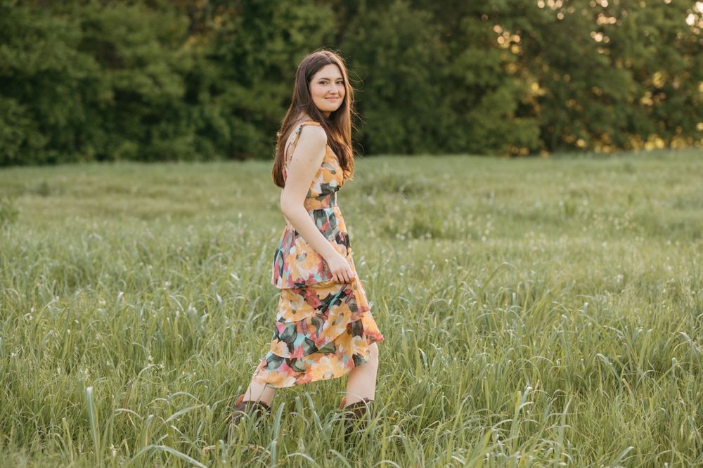 Woman in colorful floral midi dress walking through green grass field with trees in background.