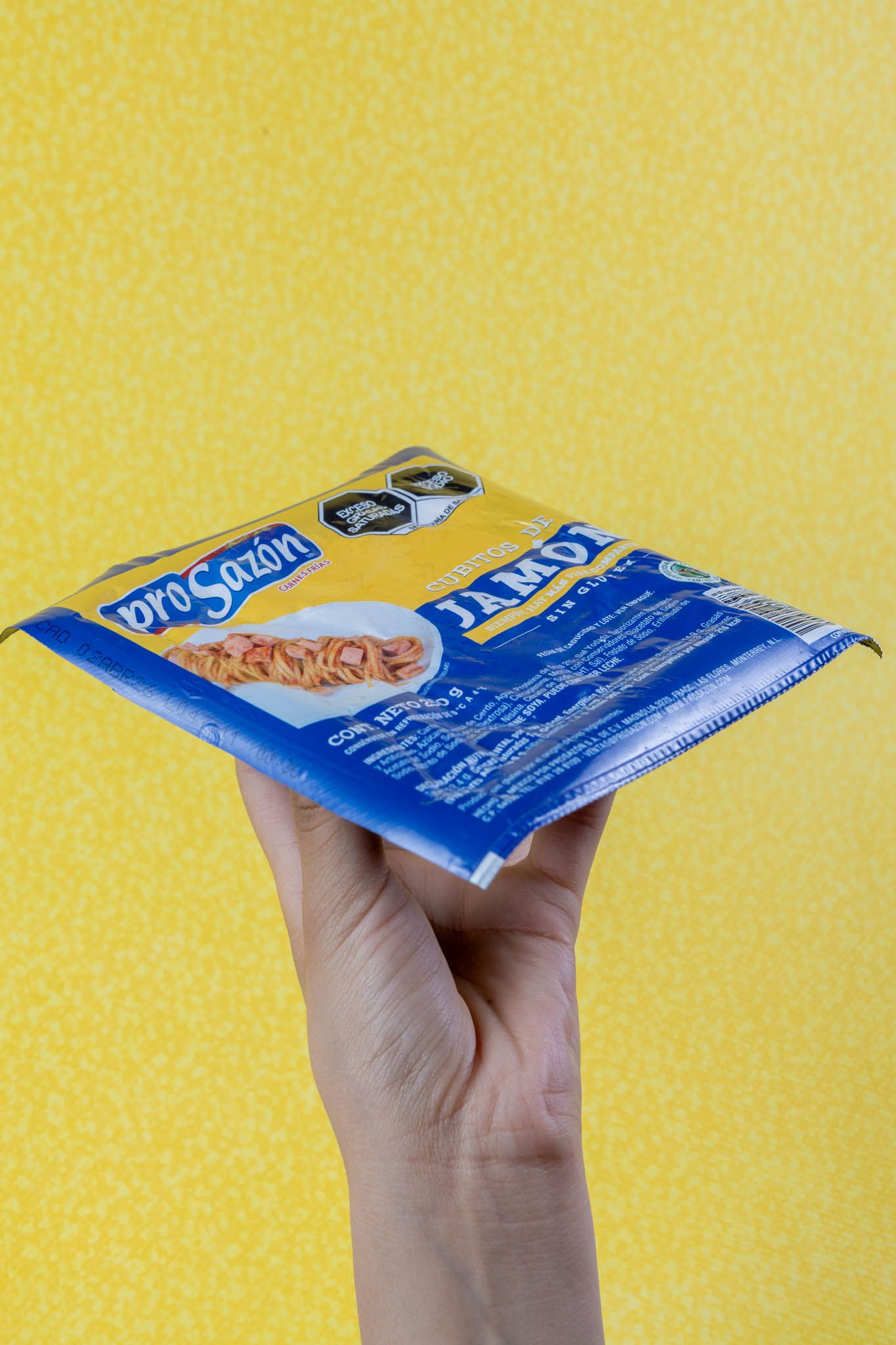 Hand holding a blue AroSazon Jamon seasoning packet against a bright yellow background.