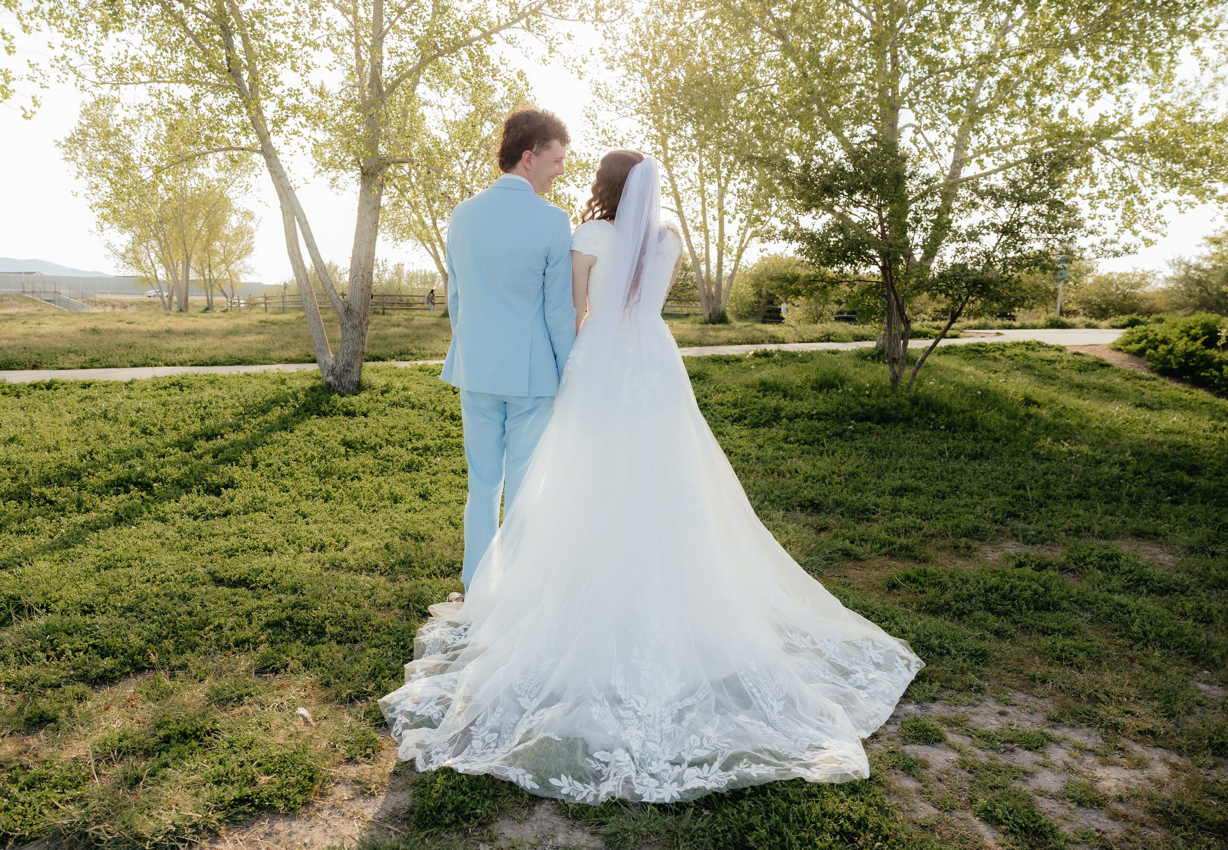 Bride in white gown with veil and groom in light blue suit walking hand in hand through a lush green park.