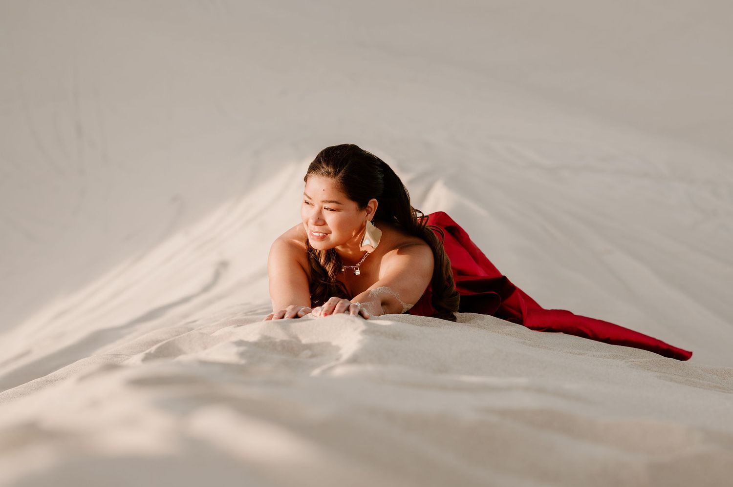 Woman in red dress lying gracefully on white sand dunes, smiling serenely in a stunning desert photoshoot.