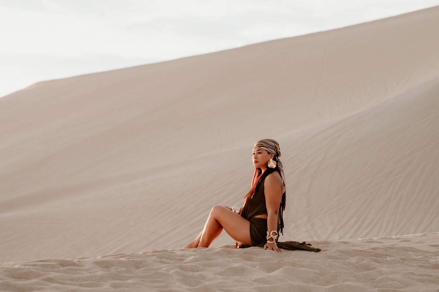 Woman in dark outfit sitting gracefully on sandy desert dunes, gazing upward in serene natural landscape.