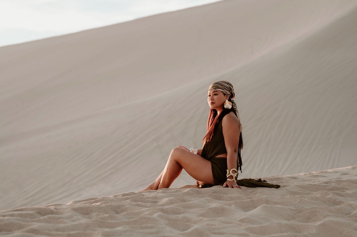 Woman in bohemian attire sitting gracefully on sandy desert dunes, wearing a head wrap and earthy tones.