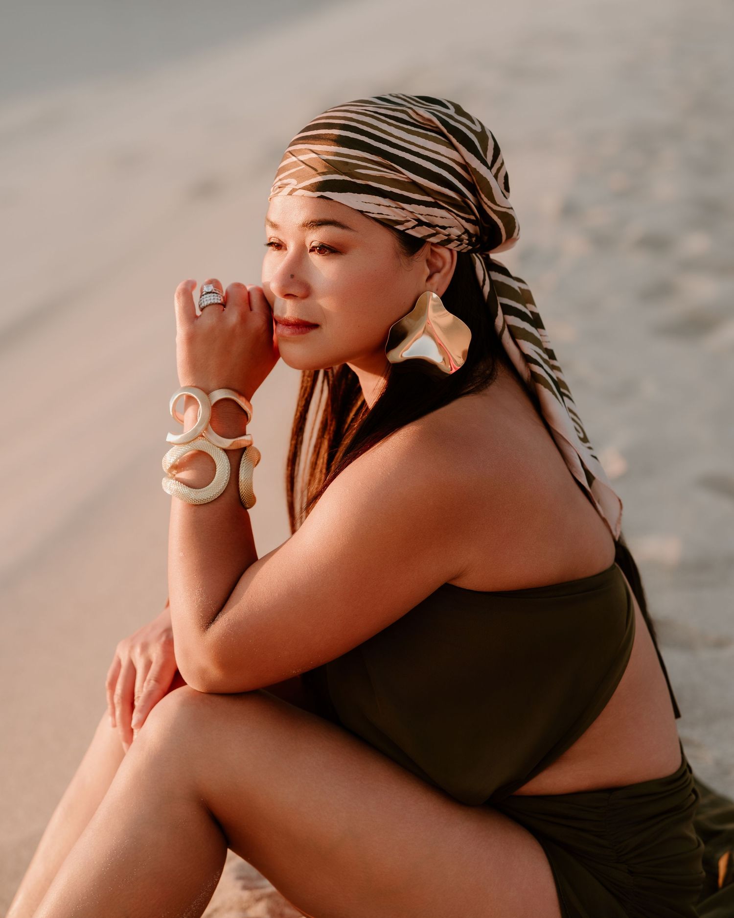 Woman in striped headwrap and bold bracelets sits on sandy beach wearing olive swimsuit in golden light.