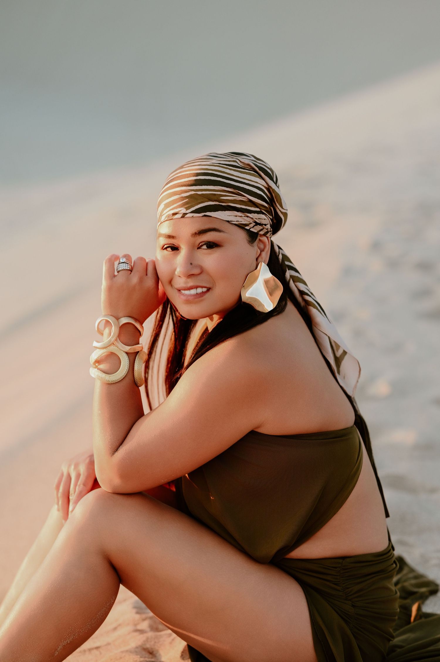Woman in olive swimsuit and patterned headscarf sits on sandy beach, wearing large hoop earrings and bangles.
