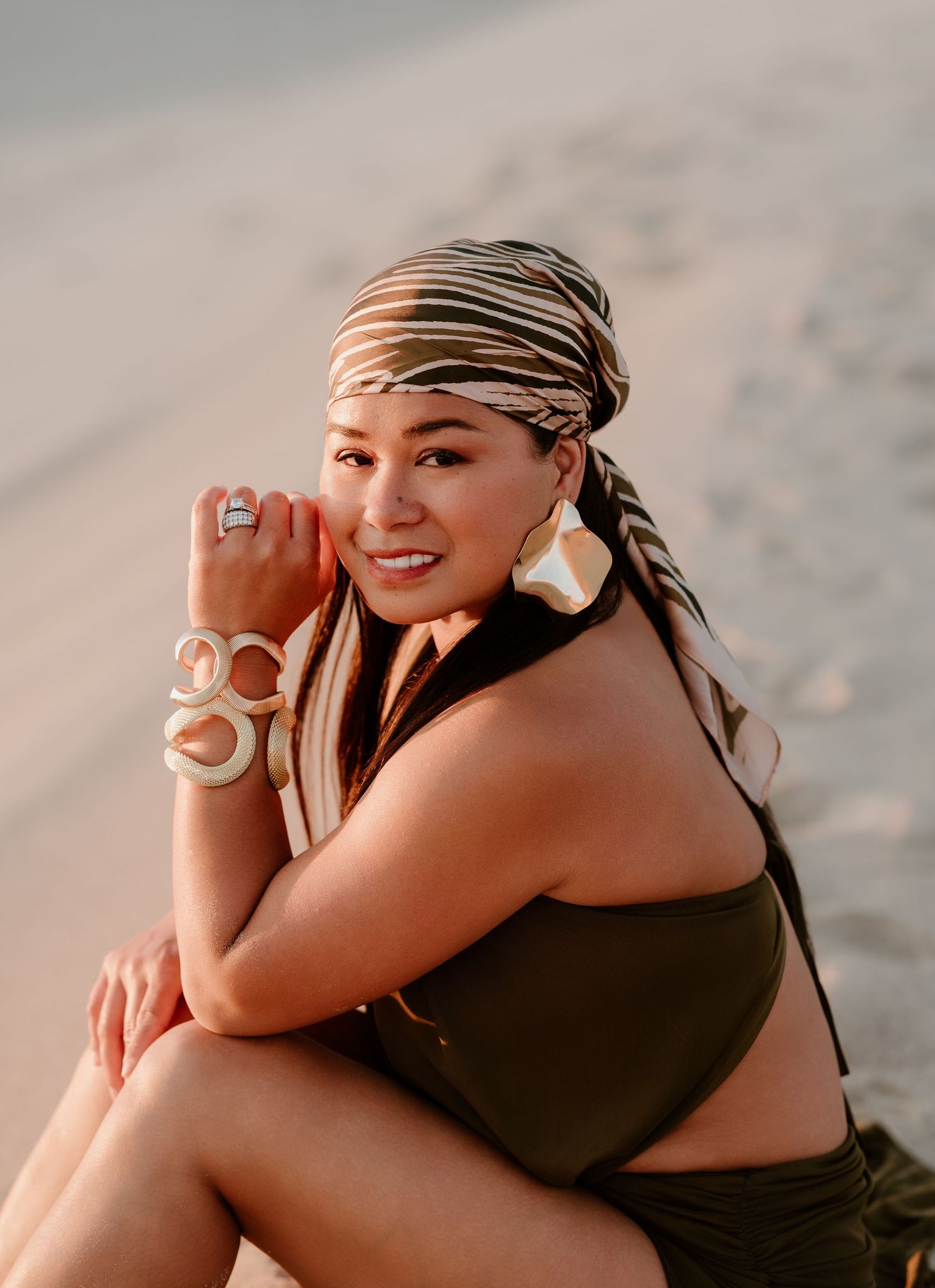 Woman in olive bikini top and striped headscarf poses on sandy beach wearing large hoop earrings and white bangles.