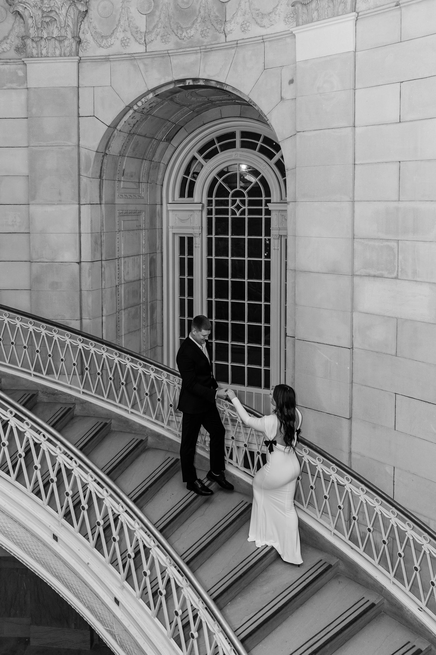 Couple in wedding attire on ornate spiral staircase of elegant stone building, black and white photography.