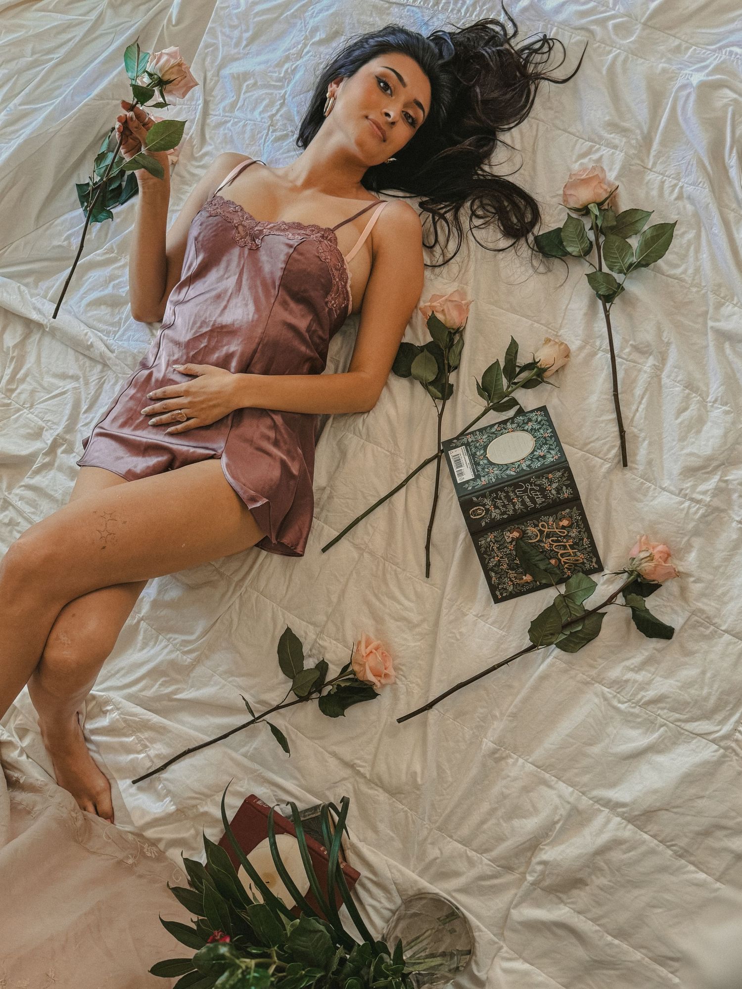 Dark-haired woman in mauve silk slip dress lying on white bed surrounded by pink roses and a book.