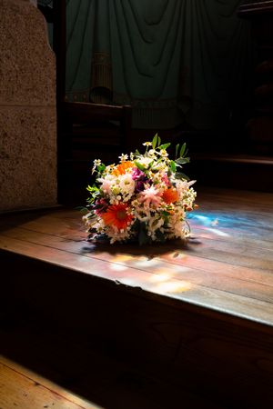 Bouquet de mariage coloré composé de gerberas orange et de fleurs blanches, posé sur le parquet d'une chapelle et éclairé par un projecteur.
