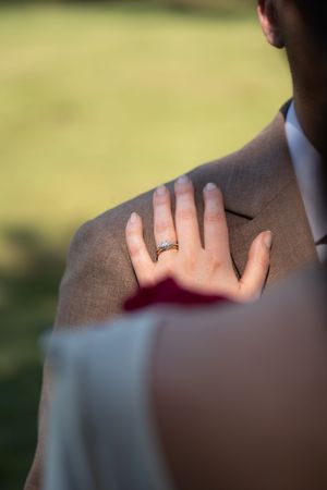La main de la mariée, où repose une alliance en diamant, posée sur l'épaule de la veste de costume beige du marié. Stéphanie Lecaudé