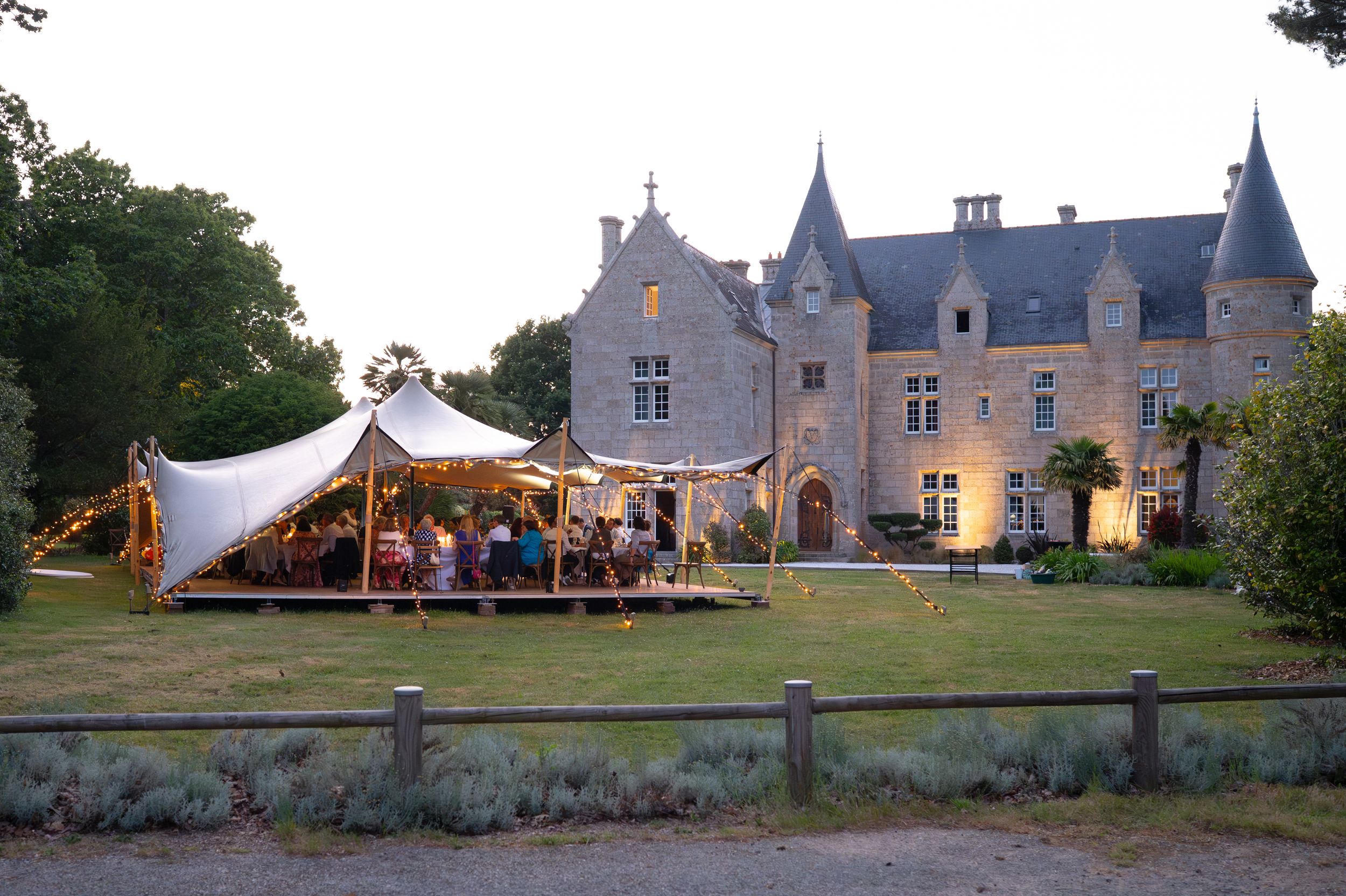 Elegant French chateau wedding venue at dusk with festive tent and guests on lawn.