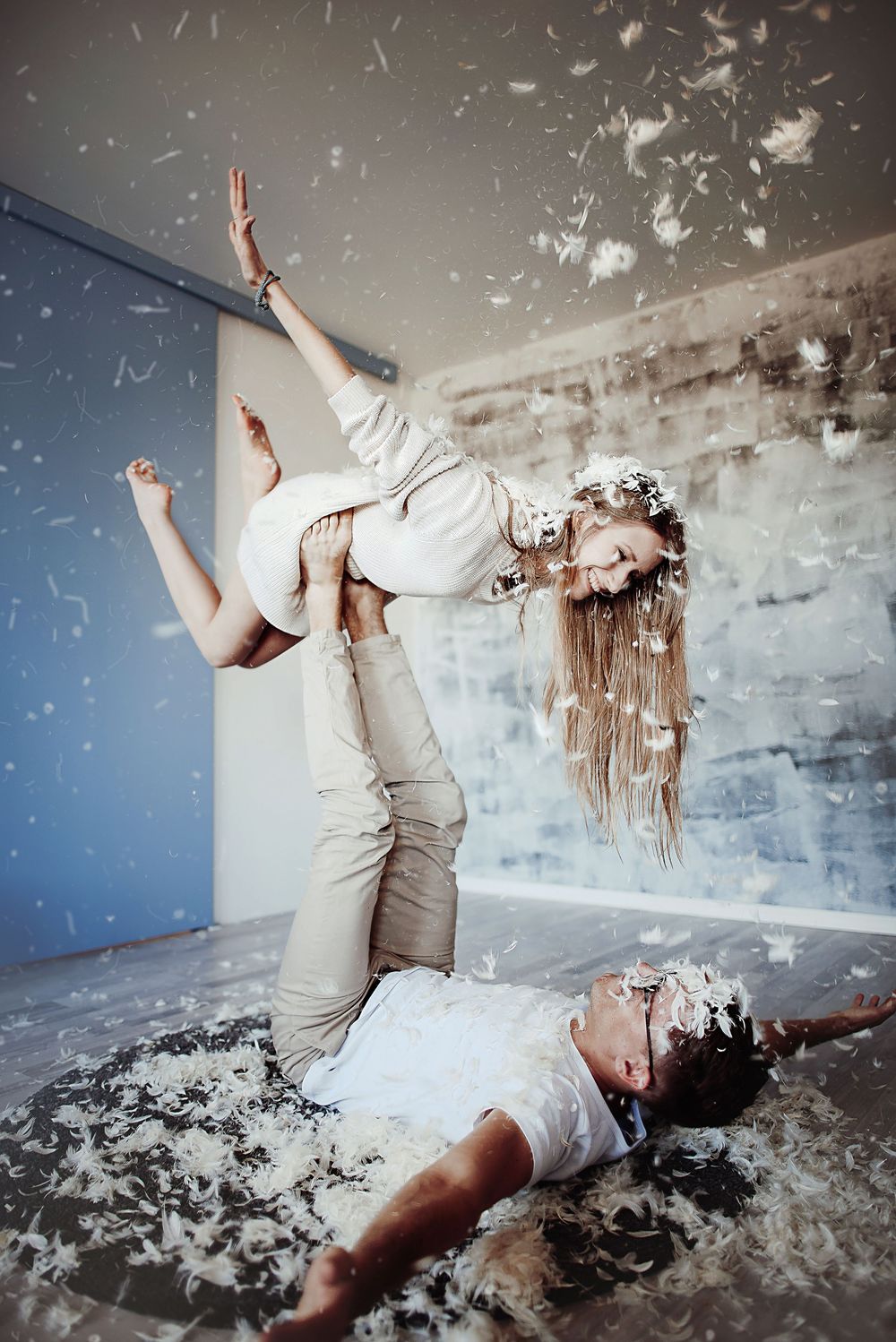 Two dancers perform an acrobatic lift surrounded by falling white feathers and confetti in a studio.