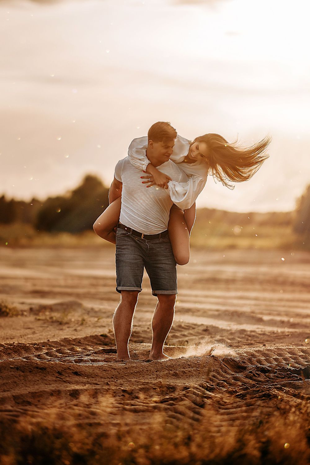 A man carries a woman over his shoulder on a sandy beach at golden sunset, her hair blowing in the wind.