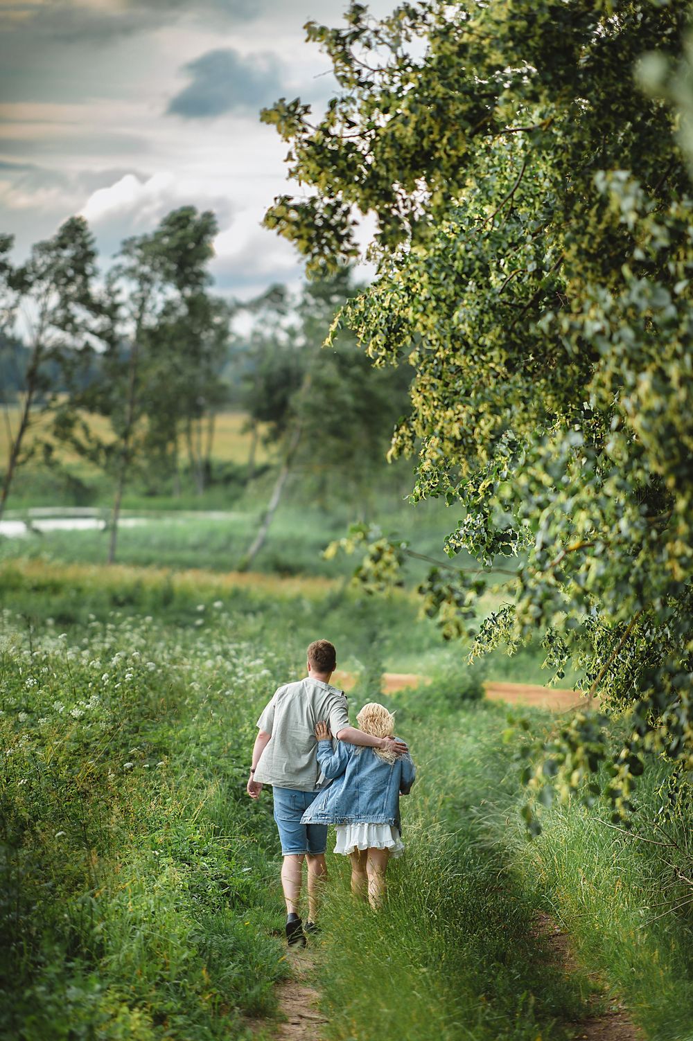 Two people walk arm in arm along a lush green countryside path lined with trees under a cloudy sky.