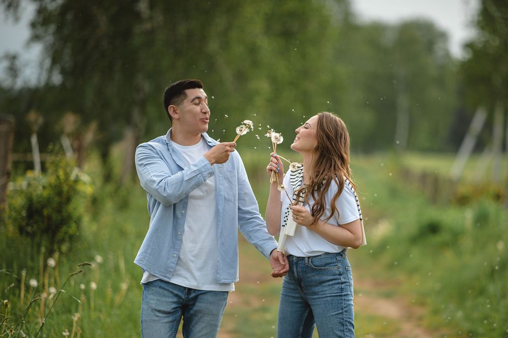 A happy couple blowing dandelion seeds together on a scenic green countryside path in summer.