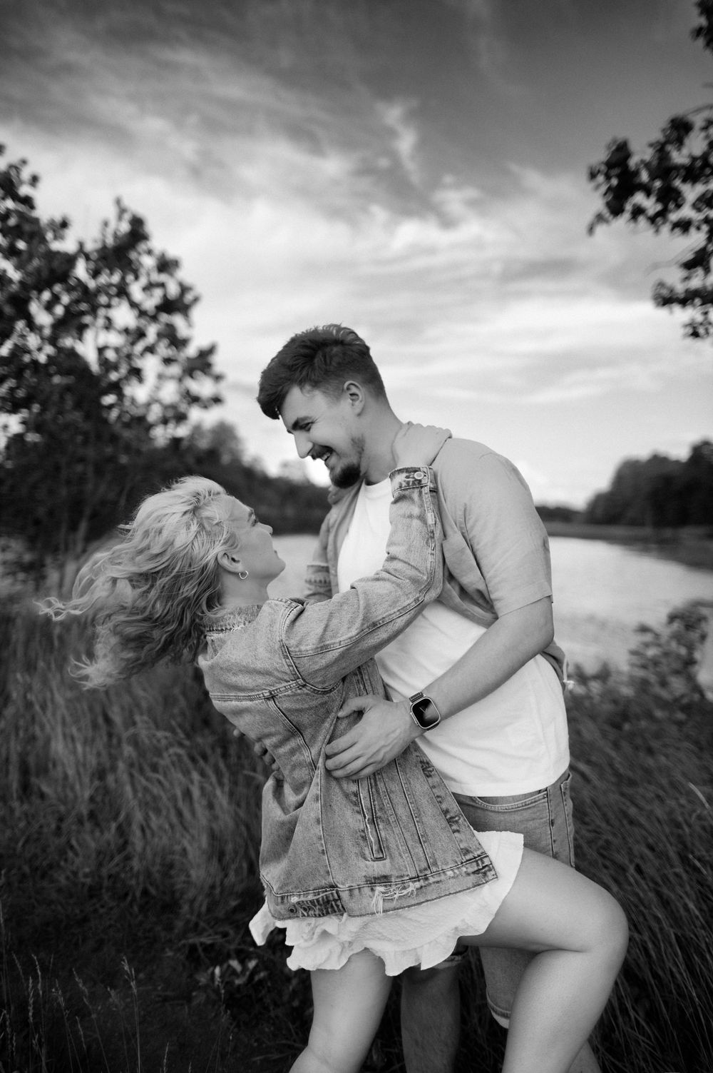 Black and white photo of a joyful couple dancing outdoors near a river, surrounded by trees and a cloudy sky.