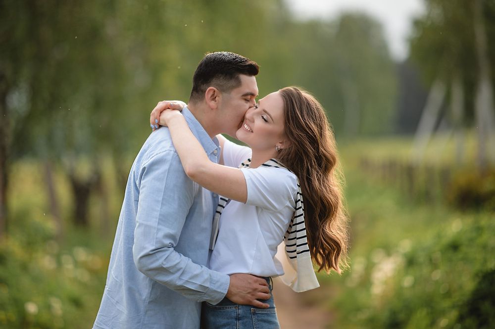 Romantic couple sharing a kiss outdoors in a lush green garden setting, woman in striped top embracing man in blue shirt.