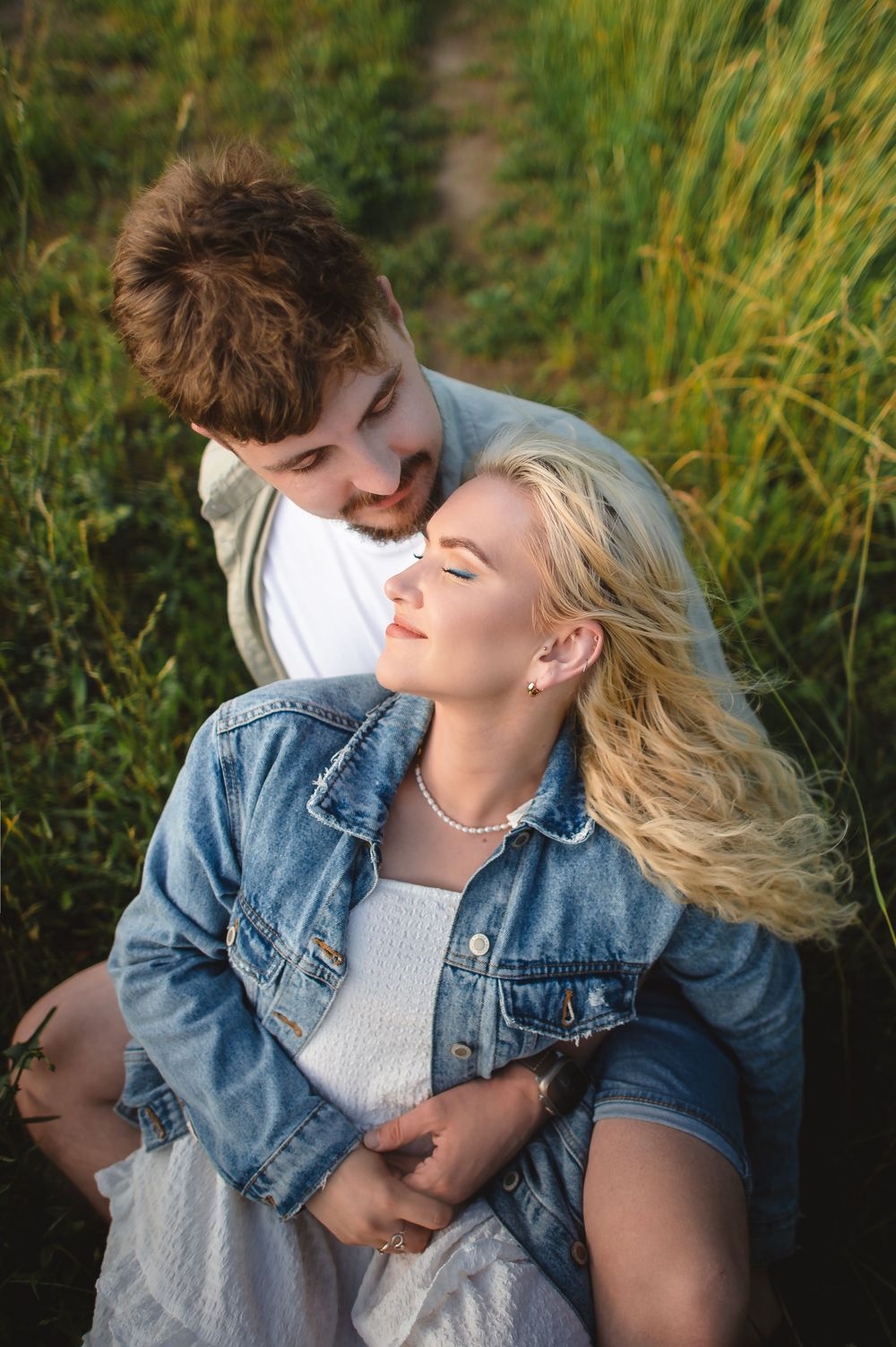A young couple shares a tender moment outdoors, with the woman leaning back into the man's embrace near lush green grass.