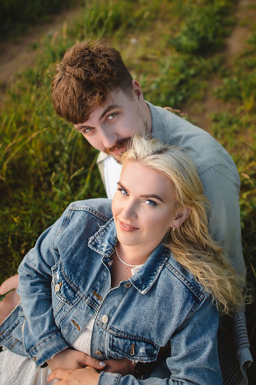 A young couple sitting in a grassy field, woman in denim jacket with blonde hair, man with mustache embracing her.