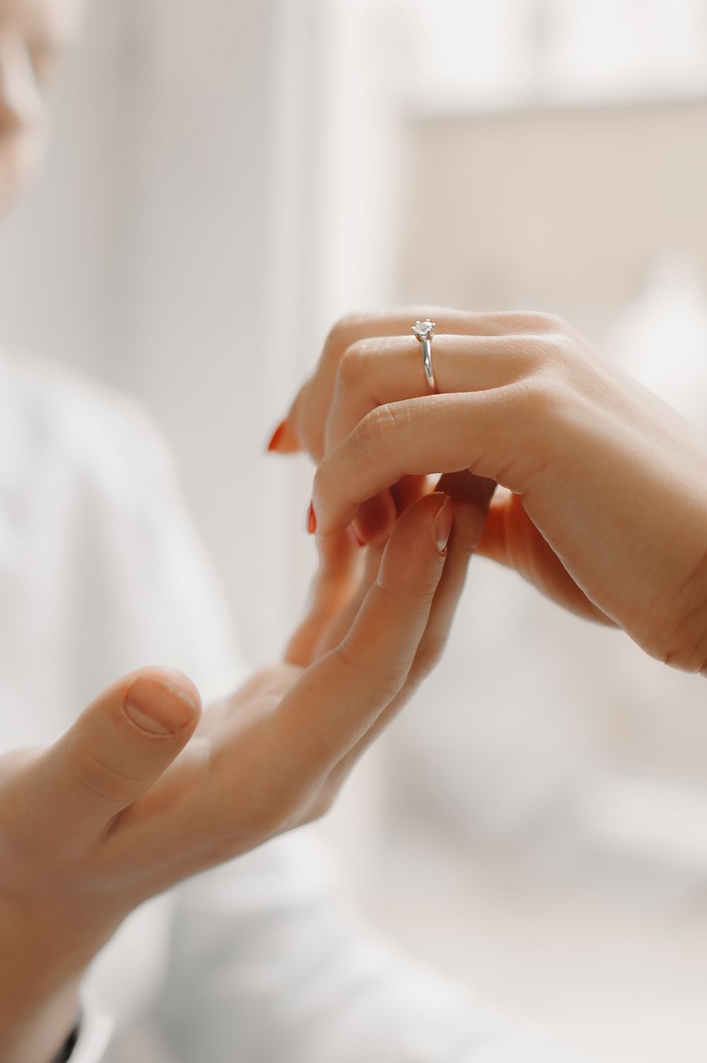 Close-up of a person placing a diamond solitaire engagement ring on a woman's finger with red nails.
