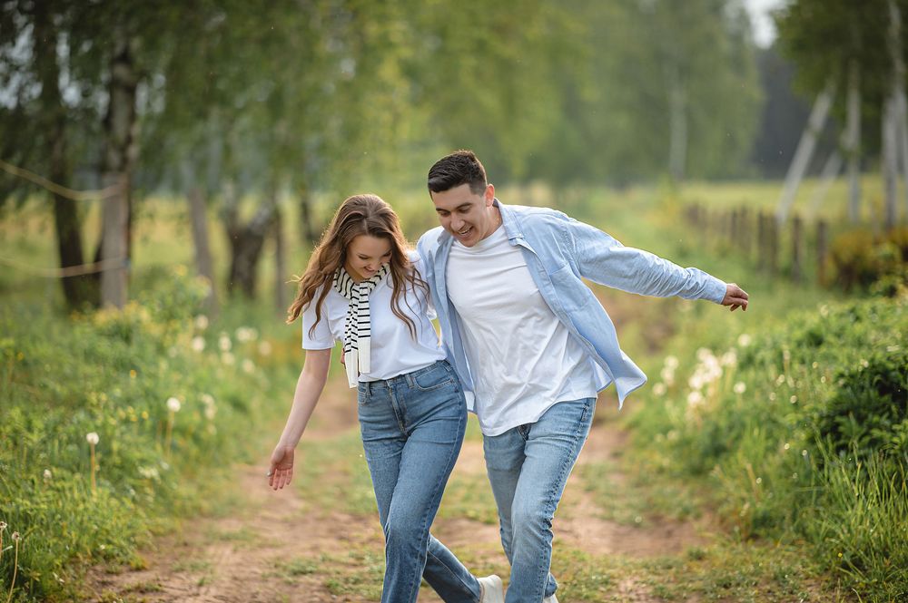 Young couple laughing and walking together on a scenic rural path surrounded by lush green trees.