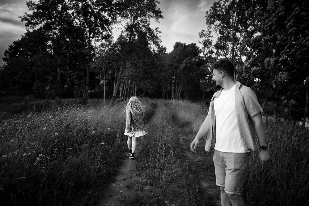 Black and white photo of a man and young girl walking on a forest path surrounded by tall grass and trees.