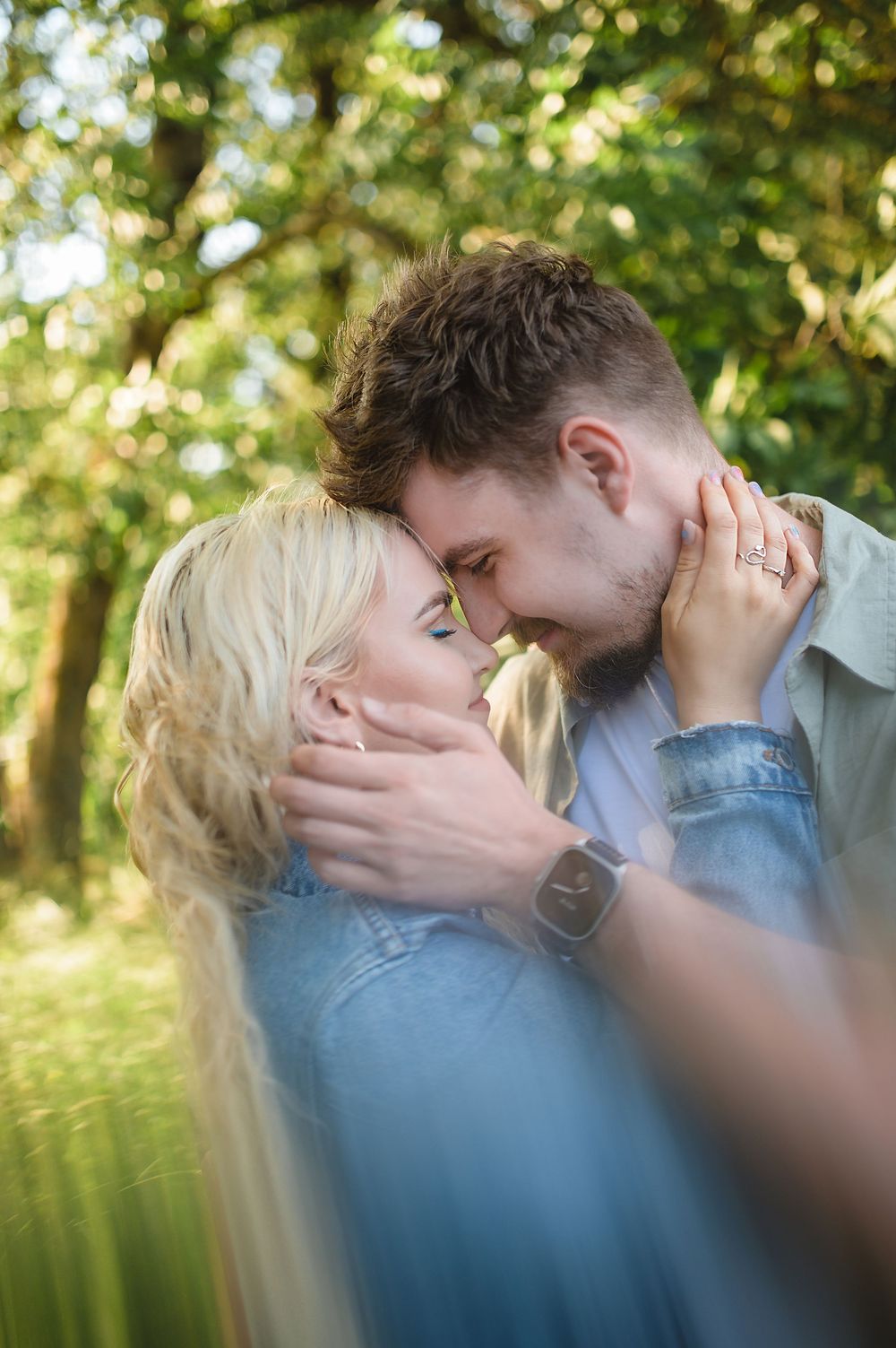A romantic couple sharing an intimate moment outdoors, touching foreheads in a lush green park setting.