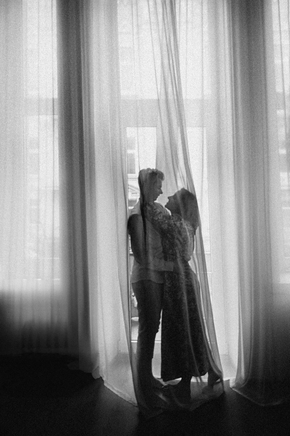 Black and white silhouette of a romantic couple embracing by sheer curtains near a bright window.