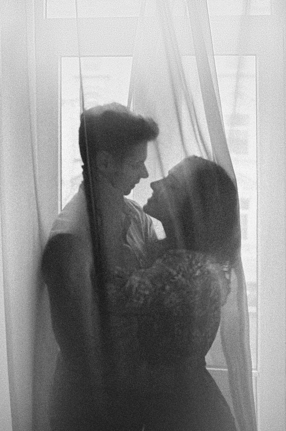 Black and white romantic couple silhouette embracing by sheer curtained window in soft natural light.